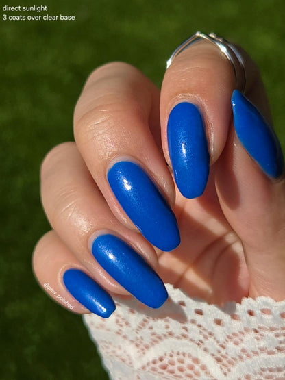 Close-up of a hand with glossy bright blue almond-shaped nails, wearing a silver ring, against a blurred green grass background. Text in the top-left corner reads 'direct sunlight 3 coats over clear base'.