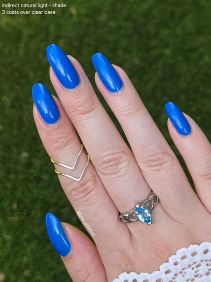 A close-up of a hand with long, glossy cobalt blue manicured nails, wearing a silver chevron ring and a silver knot ring with a blue stone, set against a blurred green grass background. Text in the top-left corner reads 'indirect natural light - shade 3 coats over clear base'.