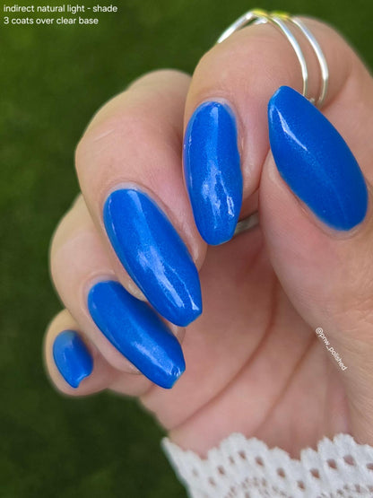 A close-up of a hand with long, almond-shaped nails painted in a glossy, vibrant royal blue shade. The nails are shown against a blurred green background, with a white lace cuff visible at the wrist and silver rings on the fingers.