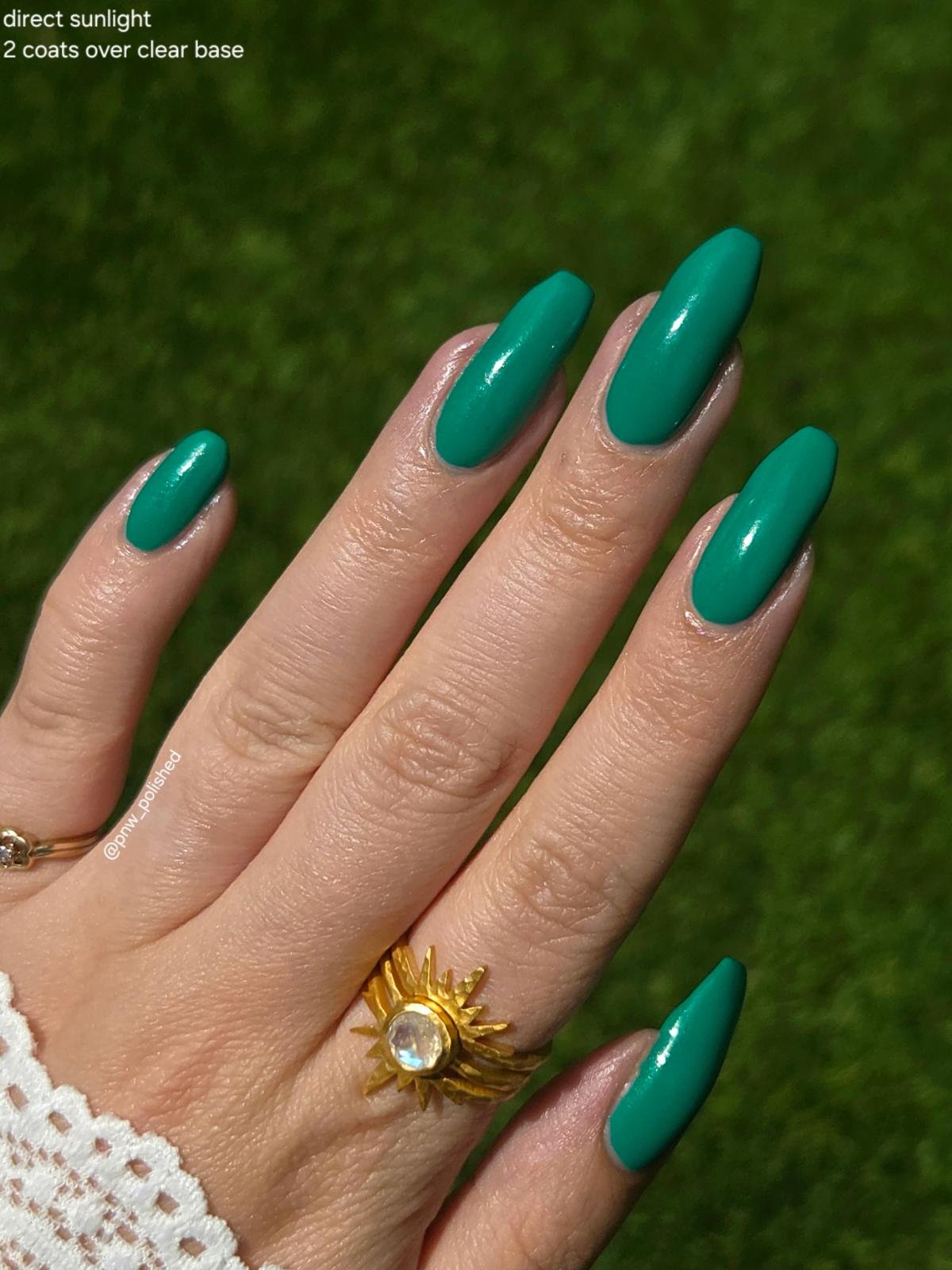 A close-up of a hand with long, almond-shaped nails painted in a glossy teal green color, wearing a gold sunburst ring, set against a blurred green grass background. Text in the top-left corner reads 'direct sunlight 2 coats over clear base'.