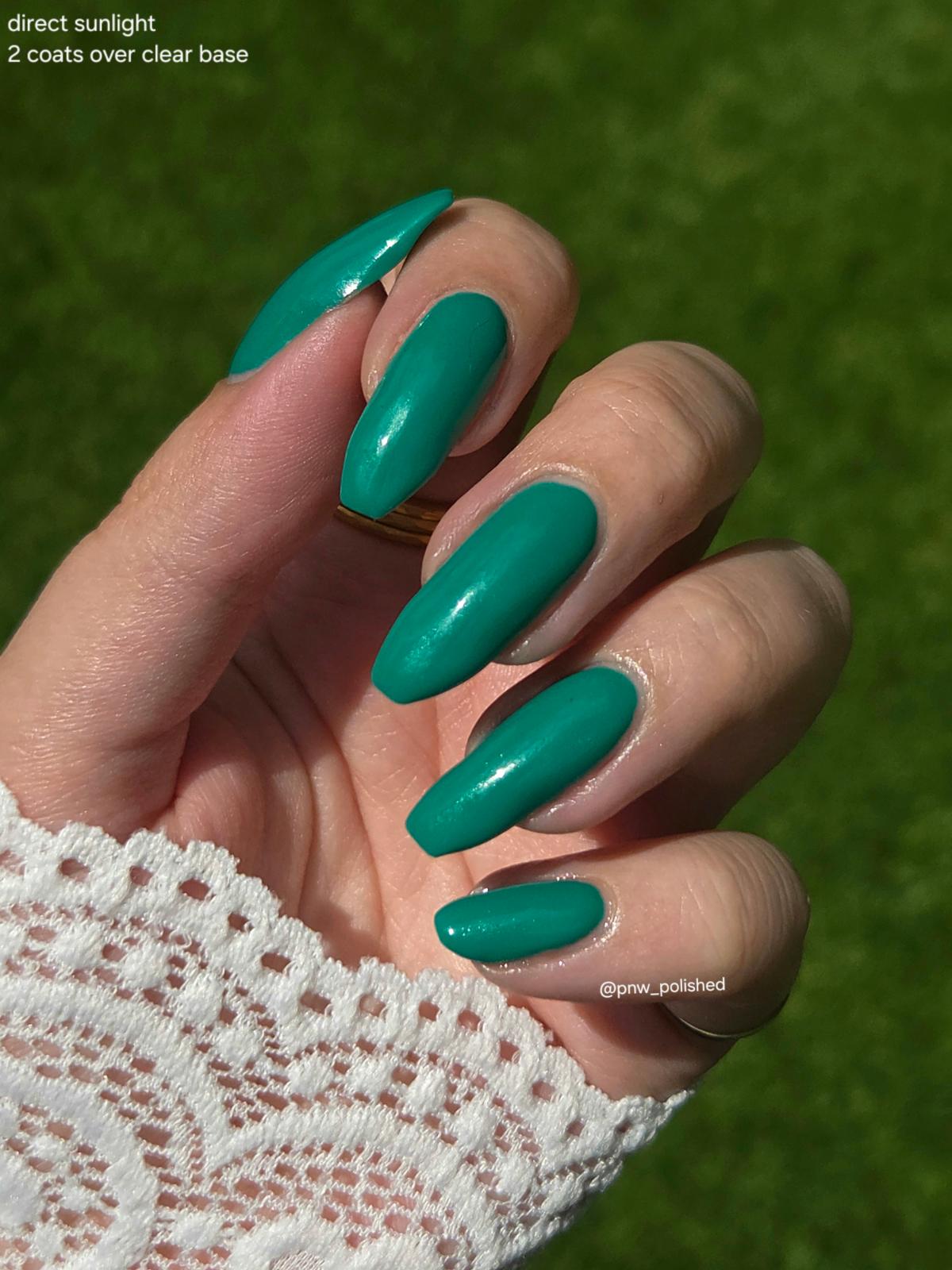 A close-up of a hand with almond-shaped nails painted in a glossy teal green color, shown outdoors against a blurred green grass background. White lace is visible on the wrist, and text in the top-left corner reads 'direct sunlight 2 coats over clear base'.