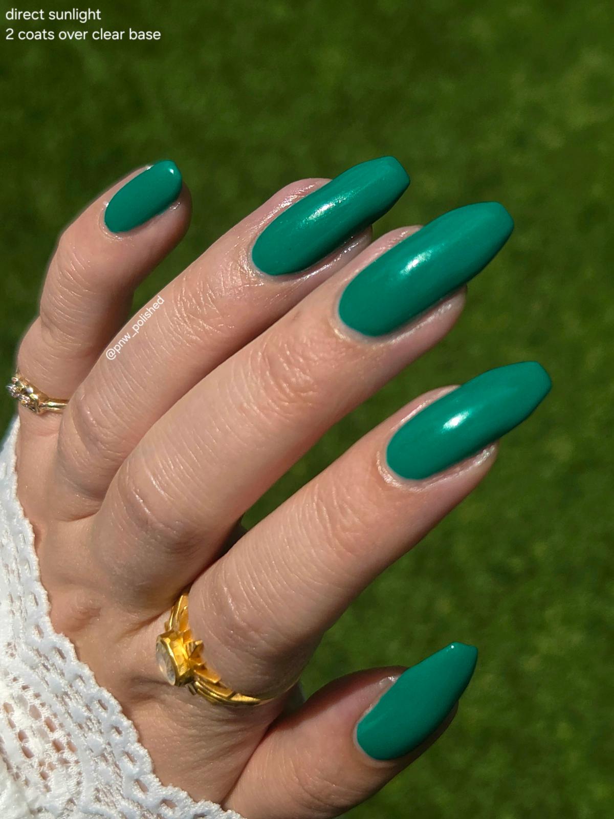 A close-up of a hand with long almond-shaped nails painted in a glossy teal green color, wearing gold rings and a white lace sleeve, set against a green grass background. Text in the top-left corner reads 'direct sunlight 2 coats over clear base'.