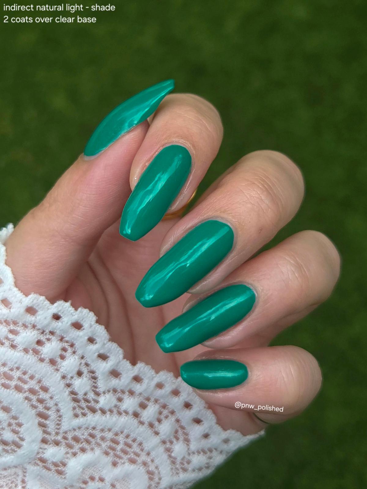 A close-up of a hand with long, almond-shaped nails painted in a glossy metallic teal green shade, wearing a white lace sleeve against a blurred green outdoor background. Text in the top-left corner reads 'indirect natural light - shade 2 coats over clear base'.