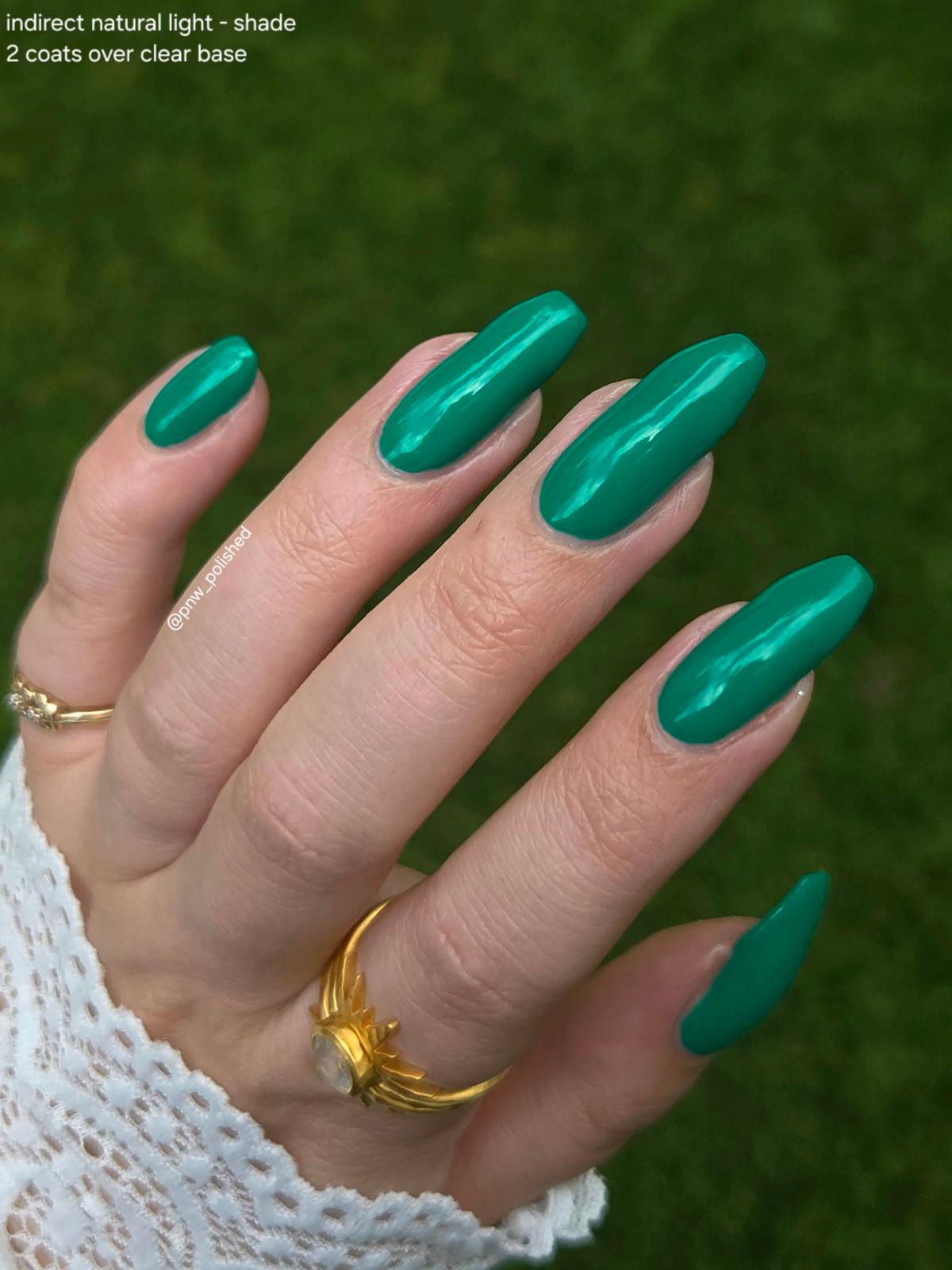 A close-up of a hand with long, glossy emerald green press-on nails, wearing gold rings and a white lace sleeve, set against a blurred green grass background. Text in the top-left corner reads 'indirect natural light - shade 2 coats over clear base'.