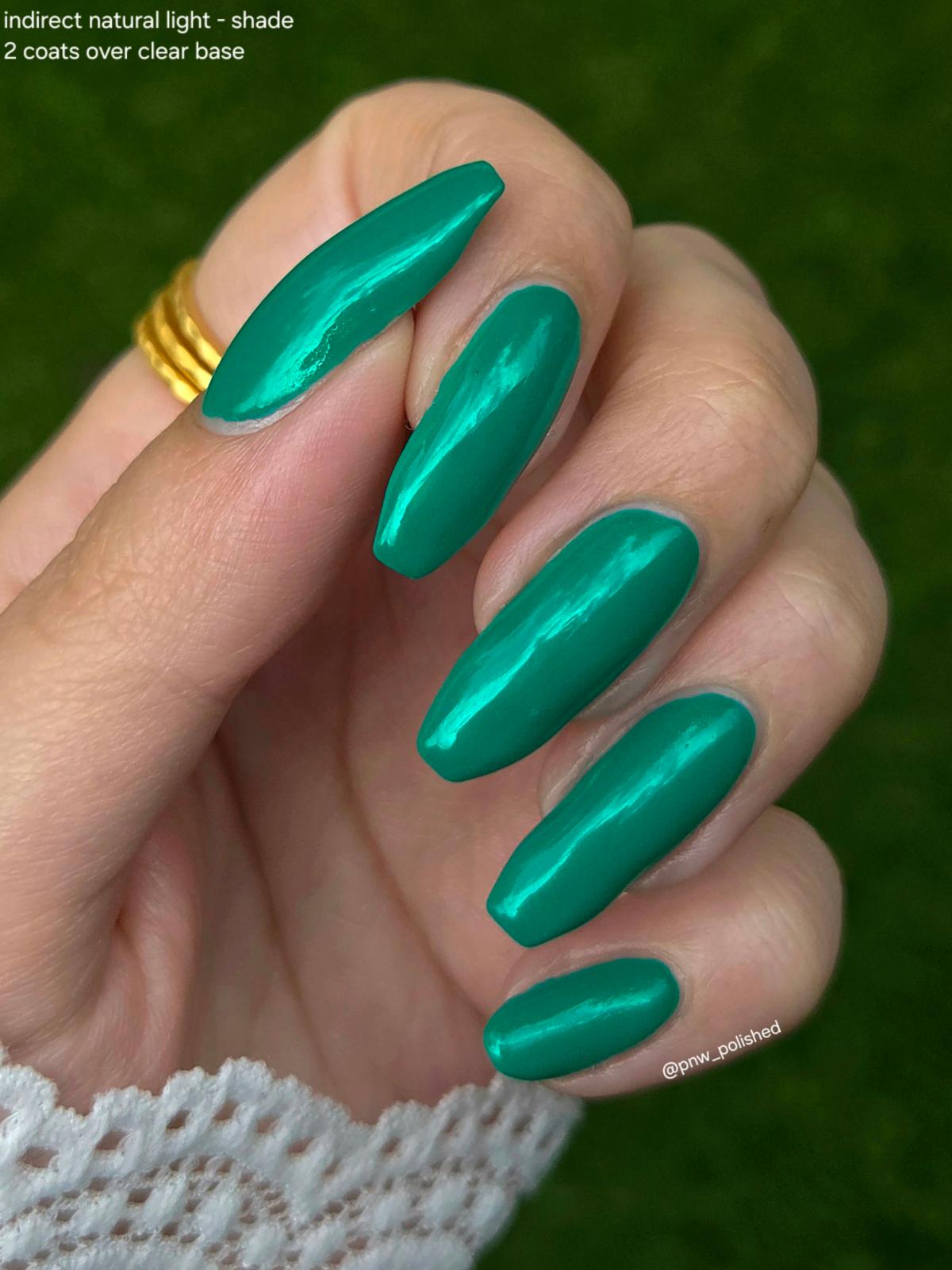 A close-up of a hand with long, almond-shaped nails painted in a glossy metallic teal-green shade, with a white lace cuff and a gold ring visible against a blurred green outdoor background.