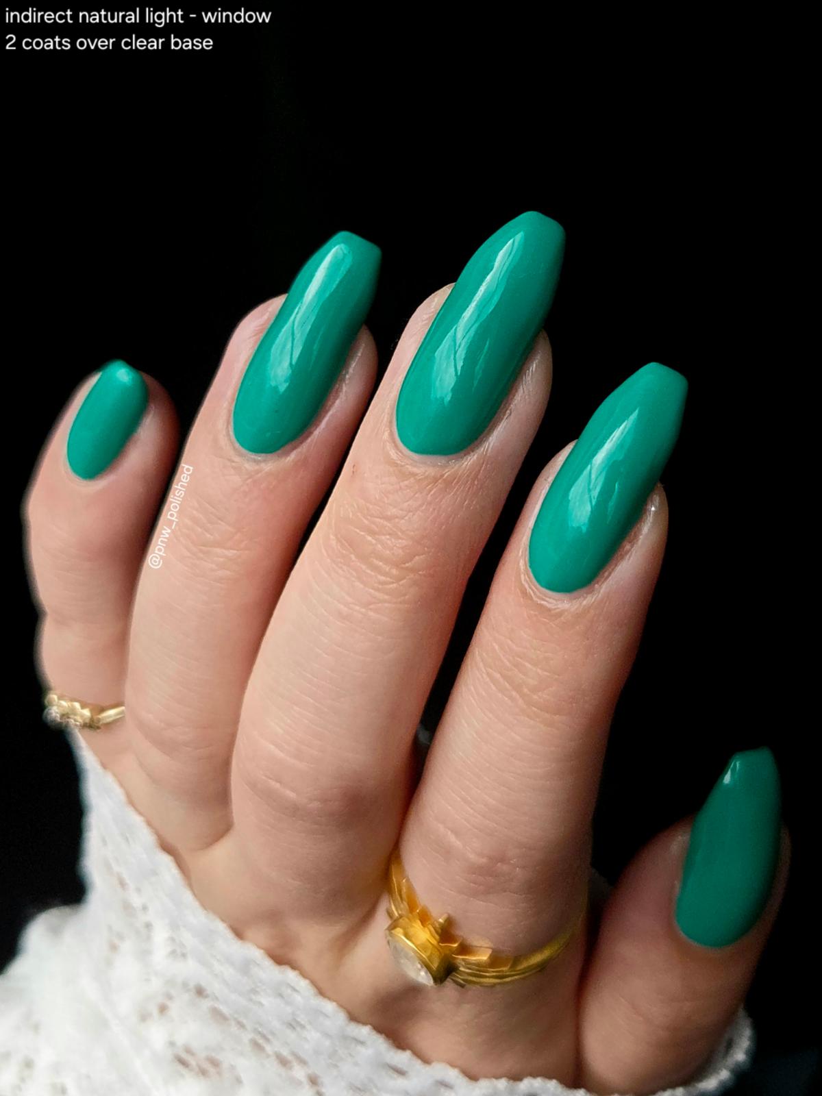 A close-up of a hand with almond-shaped nails painted in a glossy teal green color, wearing gold rings and a white lace sleeve against a black background. Text in the top-left corner reads 'indirect natural light - window 2 coats over clear base'.