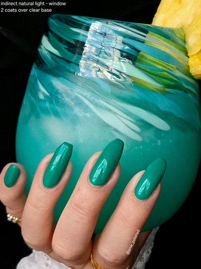 A close-up of a hand with long, glossy teal-green press-on nails holding a turquoise drink with a pineapple garnish. Text in the top-left corner reads 'indirect natural light - window 2 coats over clear base'.
