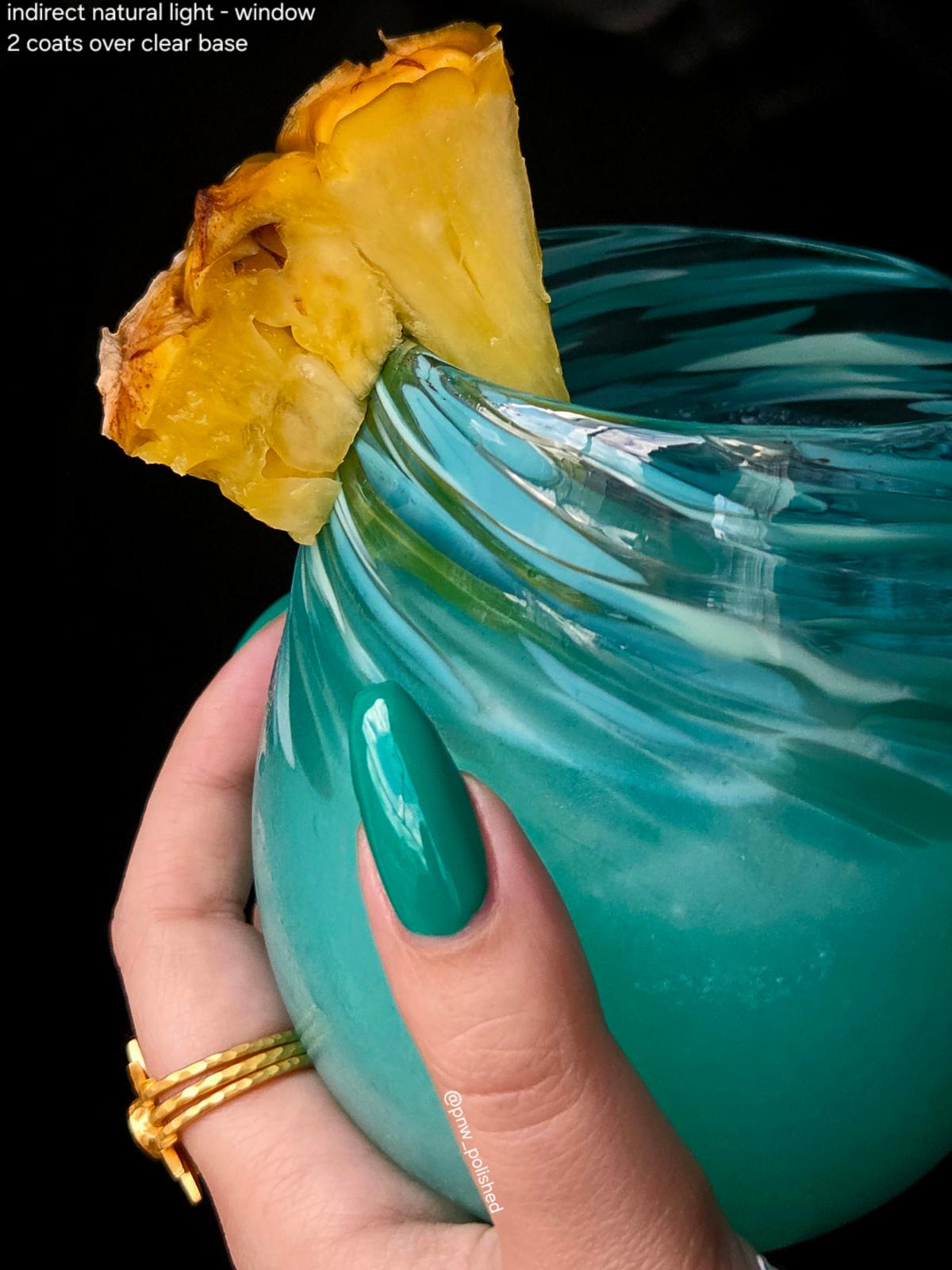 A close-up of a hand holding a turquoise blue glass tumbler with a pineapple wedge garnish, against a black background. The hand has teal nail polish and gold rings.