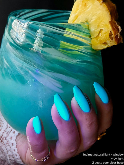 A close-up of a hand with long, bright turquoise-blue manicured nails holding a swirled clear glass filled with blue-green cocktail liquid and garnished with pineapple, against a dark background. Small text at the bottom right describes the nail finish.