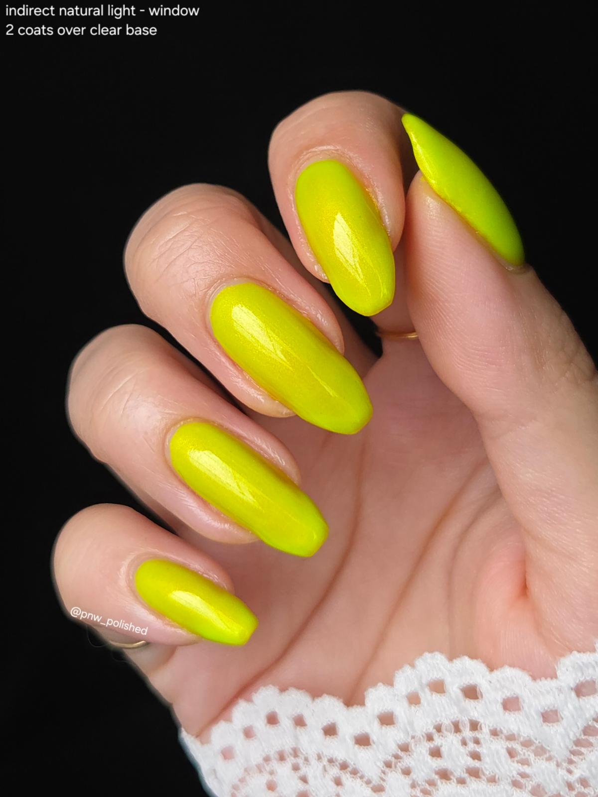 A close-up of a hand with long, almond-shaped nails painted in a bright neon yellow-green polish, shown against a black background with a small text overlay in the top left and a white lace cuff at the bottom.