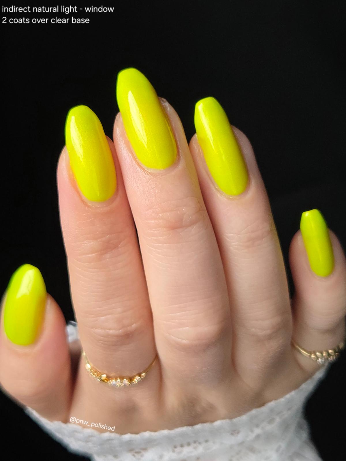 A close-up of a hand with long, almond-shaped nails painted in a bright neon yellow polish, with text in the top-left corner reading 'indirect natural light - window 2 coats over clear base'.