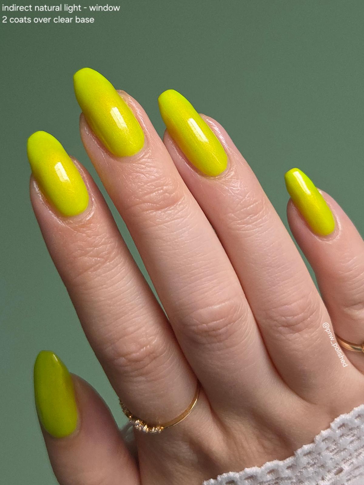 A close-up of a hand with long almond-shaped nails painted in a bright neon yellow polish, set against a solid muted green background. Small text in the top-left corner reads 'indirect natural light - window 2 coats over clear base'.