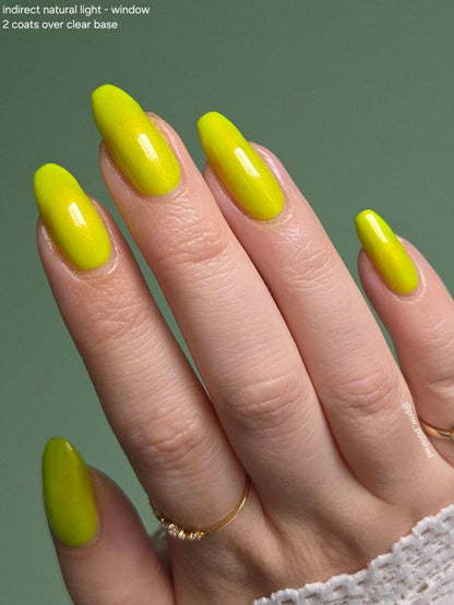 A close-up of a hand with long almond-shaped nails painted in a bright neon yellow polish, set against a solid muted green background. Small text in the top-left corner reads 'indirect natural light - window 2 coats over clear base'.