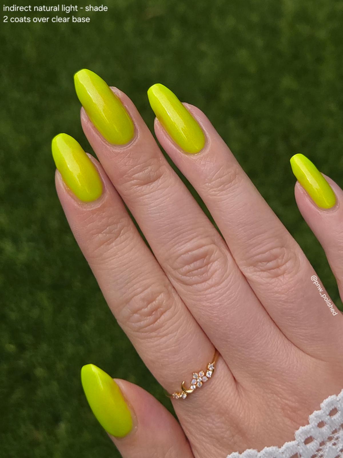 A close-up of a hand with long, almond-shaped nails painted in a bright neon yellow shade, set against a blurred green grass background. Text in the top-left corner reads 'indirect natural light - shade 2 coats over clear base'.