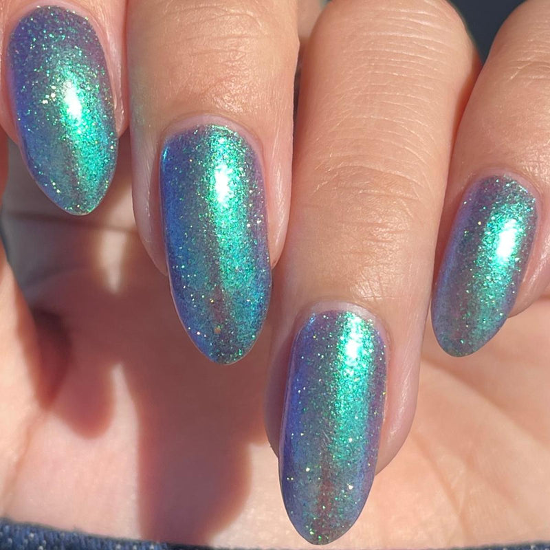 Close-up of a hand with almond-shaped nails painted in a shimmering holographic nail polish that shifts between teal, aqua, and purple with fine glitter.