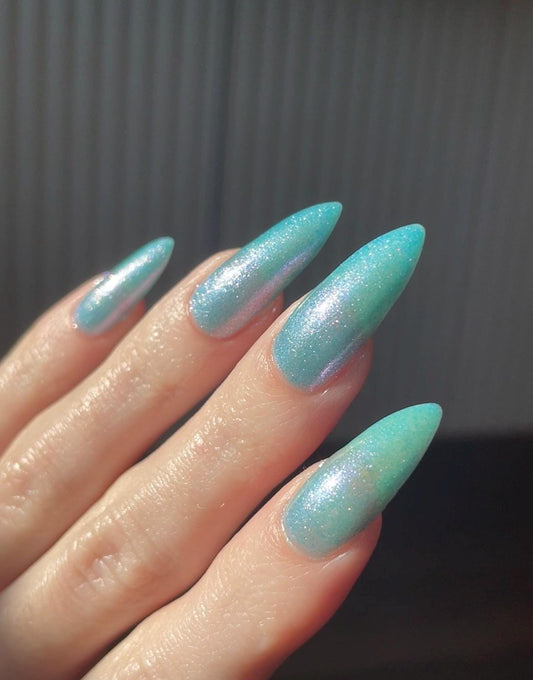 Long pale fingers end in long pointy nails painted with a pale blue shimmer nail polish. The polish has blue to pink shifting shimmer and glows in the light.