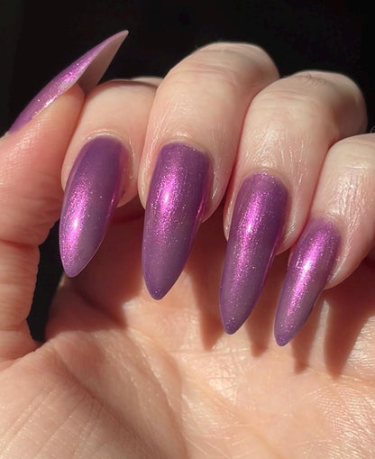 Long pale fingers end in long pointy nails painted with a medium purple shimmer nail polish. The polish has orange shimmer and scattered orange sparkle and glows in the light.