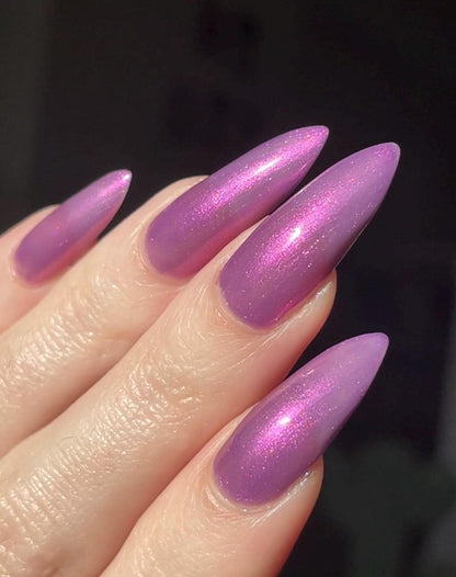 Long pale fingers end in long pointy nails painted with a medium purple shimmer nail polish. The polish has orange shimmer and scattered orange sparkle and glows in the light.