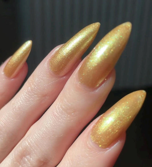 Long pale fingers end in long pointy nails painted with a golden shimmer nail polish. the polish has hints of green sparkle and glows in the light.