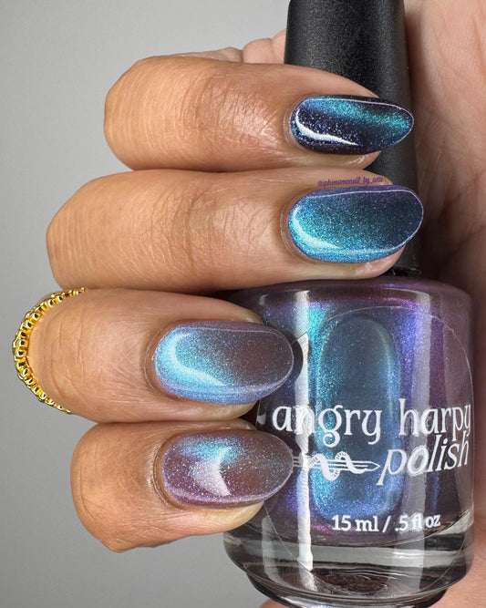 Elegant medium-brown fingers are painted with a plum polish that has a teal-blue magnetic stripe. They hold a bottle with the white Angry Hsrpy Logo on it, filled with shimmering purple-teal polish.