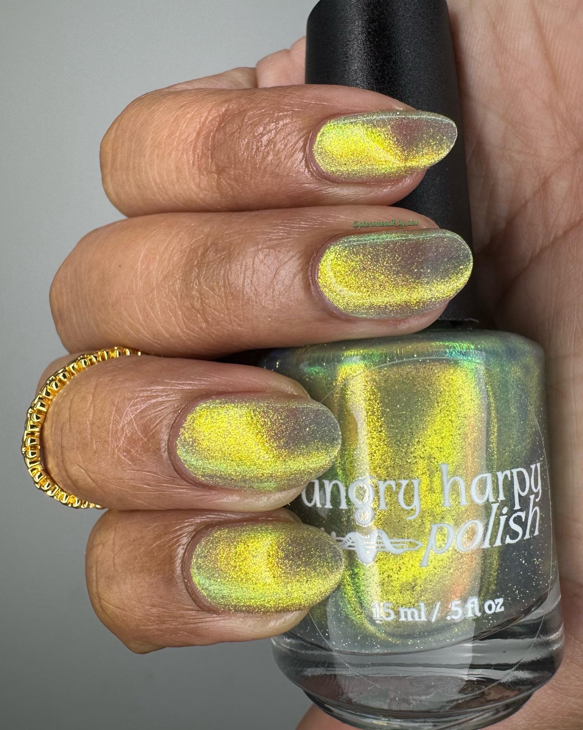 A hand with medium-dark skin holds a bottle of nail polish, showing off nails painted in a bright shifty polish that seems to glow yellow-green from within.