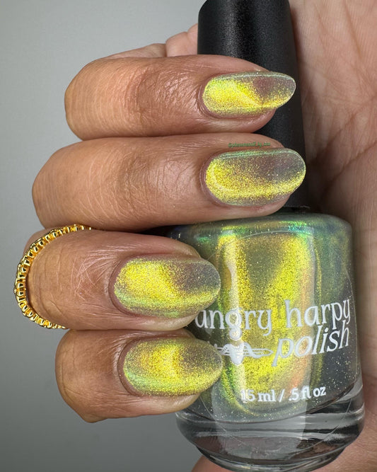 A hand with medium-dark skin holds a bottle of nail polish, showing off nails painted in a bright shifty polish that seems to glow yellow-green from within.