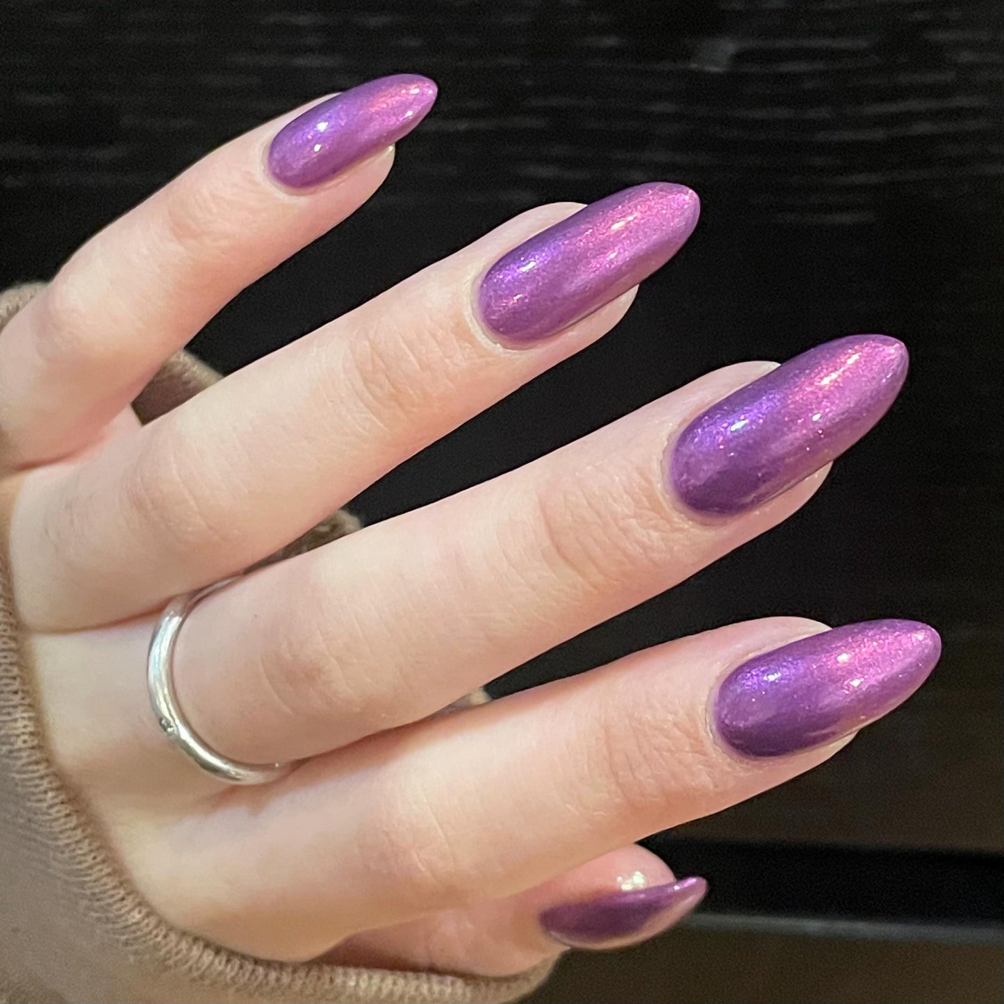 An elegant pale hand end in long oval nails painted with a medium purple shimmer nail polish. The polish has orange shimmer and scattered orange sparkle and glows in the indoor light.