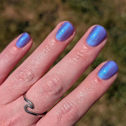 A closeup of a hand and nails displaying indigo polish with blue shimmer. Polish glows in the light.