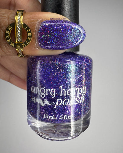 A closeup of a thumb and nail polish bottle with a vibrant violet lacquer sparkling with purple holographic glitter and green shimmer.