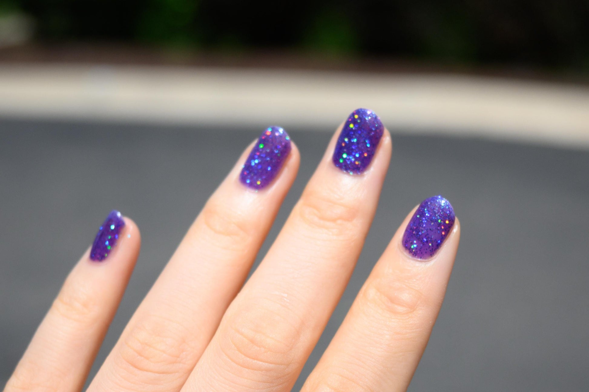 A closeup of a hand with nails painted a vibrant violet lacquer sparkling with purple holographic glitter and green shimmer.