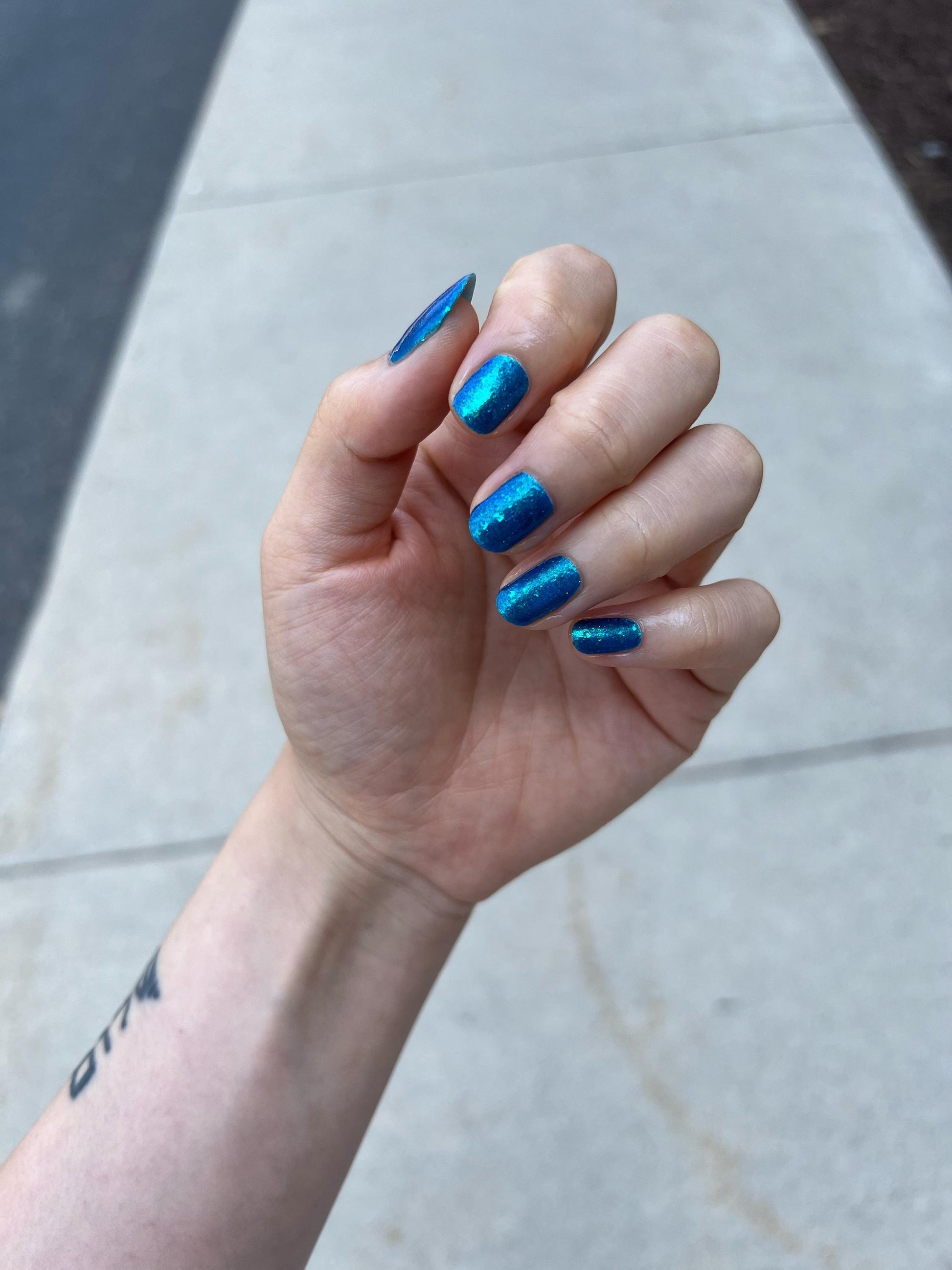 A closeup of a hand with nails painted a gorgeous, vibrant, shimmery deep teal color, packed with flakes and reflective blue glitter scattered throughout.