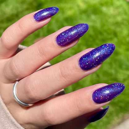A closeup of a hand with nails painted a vibrant violet lacquer sparkling with purple holographic glitter and green shimmer.