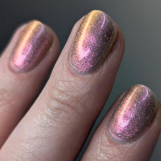 A closeup of a hand with nails painted a sheer multichrome nail polish that shifts gold-orange-pink-fuschia, with pale pink reflective glitter scattered throughout.