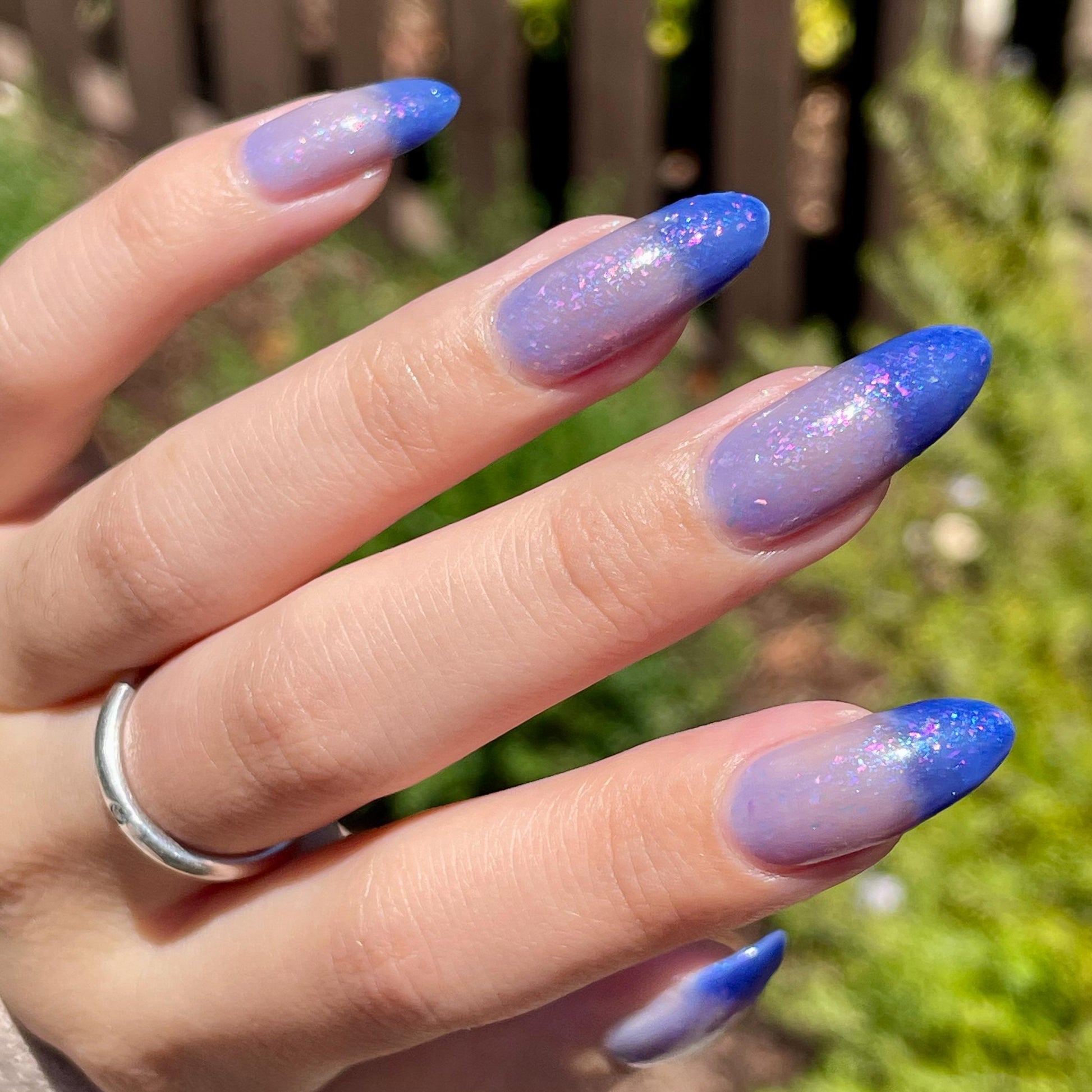 A closeup of a hand with nails painted a thermal polish that goes from dark blue at the tips (cold) to a blue-tinted clear (warm). Polish is filled with iridescent green-pink flakes and blue shimmer.