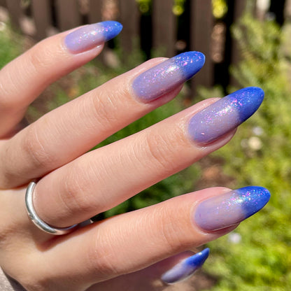 A closeup of a hand with nails painted a thermal polish that goes from dark blue at the tips (cold) to a blue-tinted clear (warm). Polish is filled with iridescent green-pink flakes and blue shimmer.