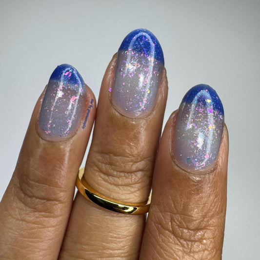A closeup of a hand with nails painted a thermal polish that goes from dark blue at the tips (cold) to a blue-tinted clear (warm). Polish is filled with iridescent green-pink flakes and blue shimmer.