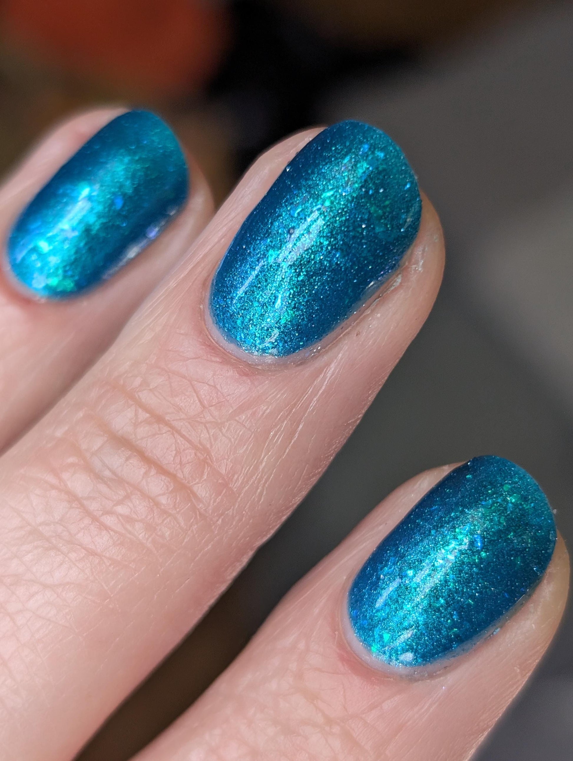 A closeup of a hand with nails painted a gorgeous, vibrant, shimmery deep teal color, packed with flakes and reflective blue glitter scattered throughout.