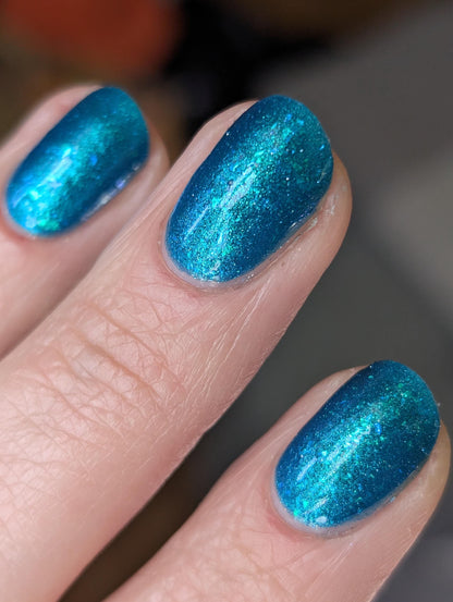 A closeup of a hand with nails painted a gorgeous, vibrant, shimmery deep teal color, packed with flakes and reflective blue glitter scattered throughout.
