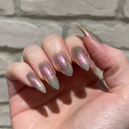 A closeup of a hand with nails painted a sheer multichrome nail polish that shifts gold-orange-pink-fuschia, with pale pink reflective glitter scattered throughout.