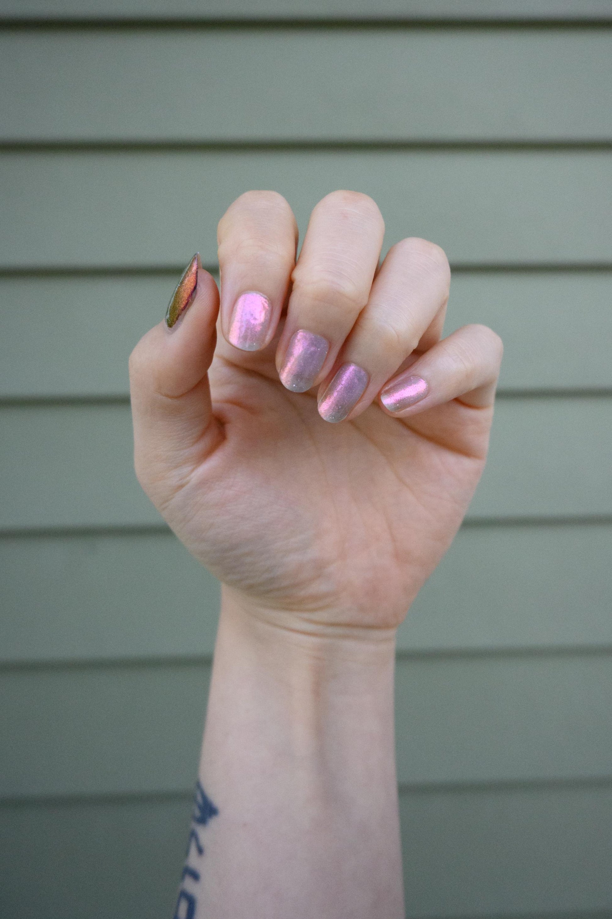 A closeup of a hand with nails painted a sheer multichrome nail polish that shifts gold-orange-pink-fuschia, with pale pink reflective glitter scattered throughout.