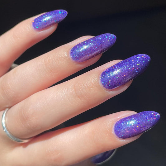 A closeup of a hand with nails painted a vibrant violet lacquer sparkling with purple holographic glitter and green shimmer.