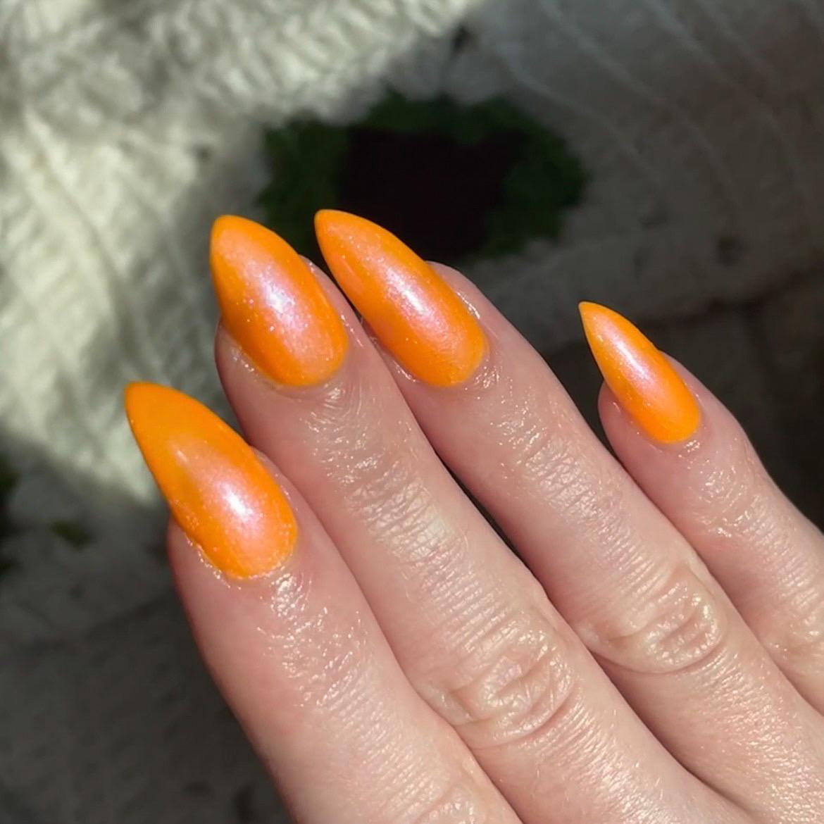 A closeup of a hand with nails painted neon orange nail lacquer with pink-purple shimmer.