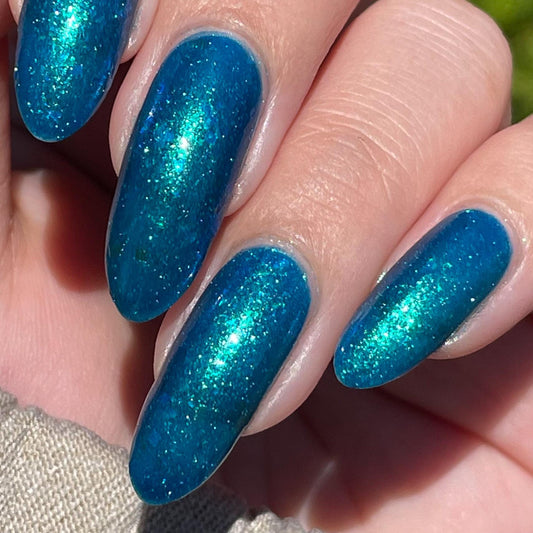 A closeup of a hand with nails painted a gorgeous, vibrant, shimmery deep teal color, packed with flakes and reflective blue glitter scattered throughout.
