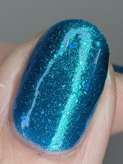 A closeup of a hand with nails painted a gorgeous, vibrant, shimmery deep teal color, packed with flakes and reflective blue glitter scattered throughout.
