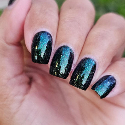 A closeup of a hand with nails painted a shiny black with bright green shimmer and pinkish reflective glitter.
