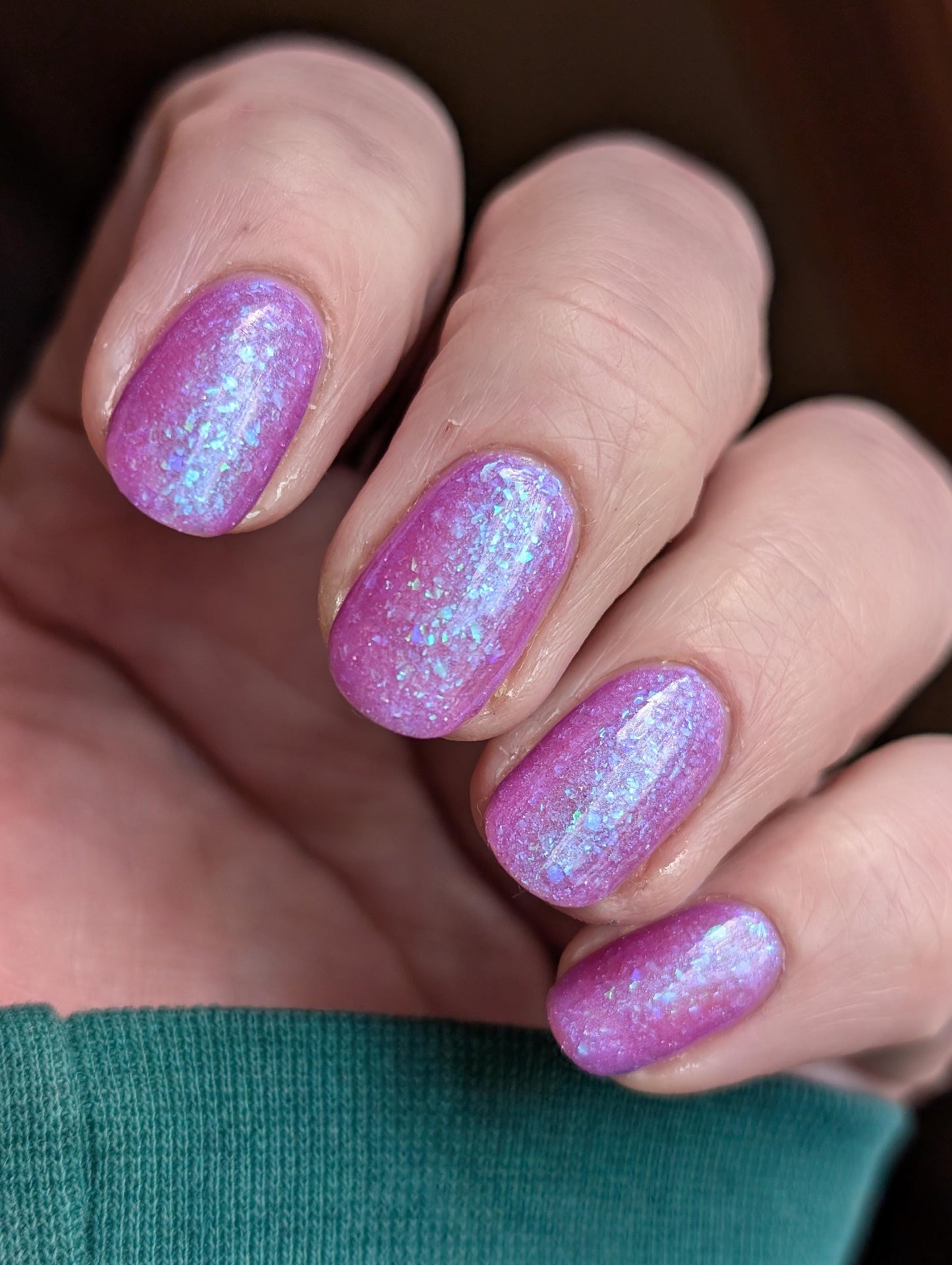 Closeup of nails painted in a lacquer that is bright pinkish purple sparkling with aqua shimmer and packed with iridescent flakes.