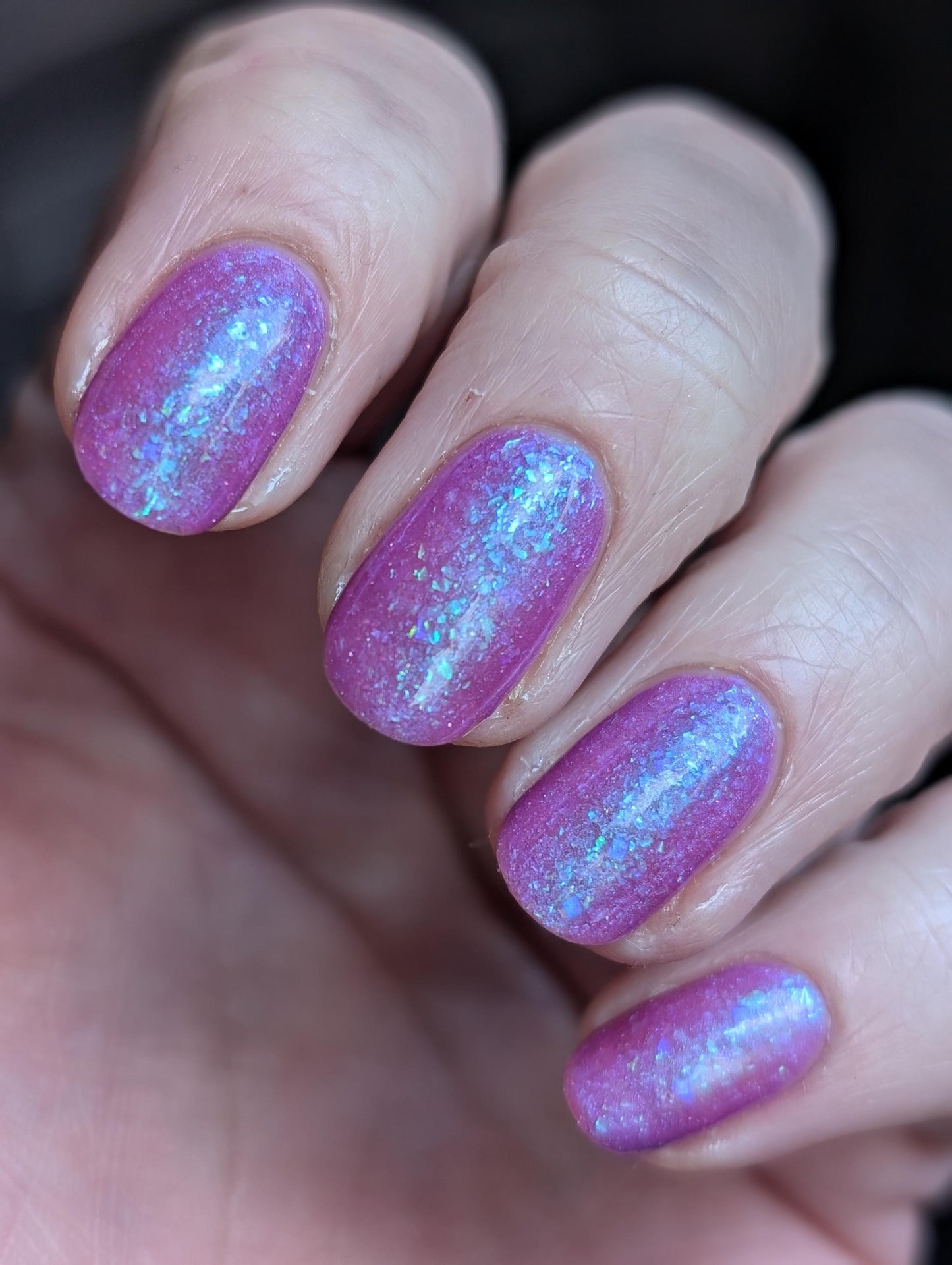 Closeup of nails painted in a lacquer that is bright pinkish purple sparkling with aqua shimmer and packed with iridescent flakes.