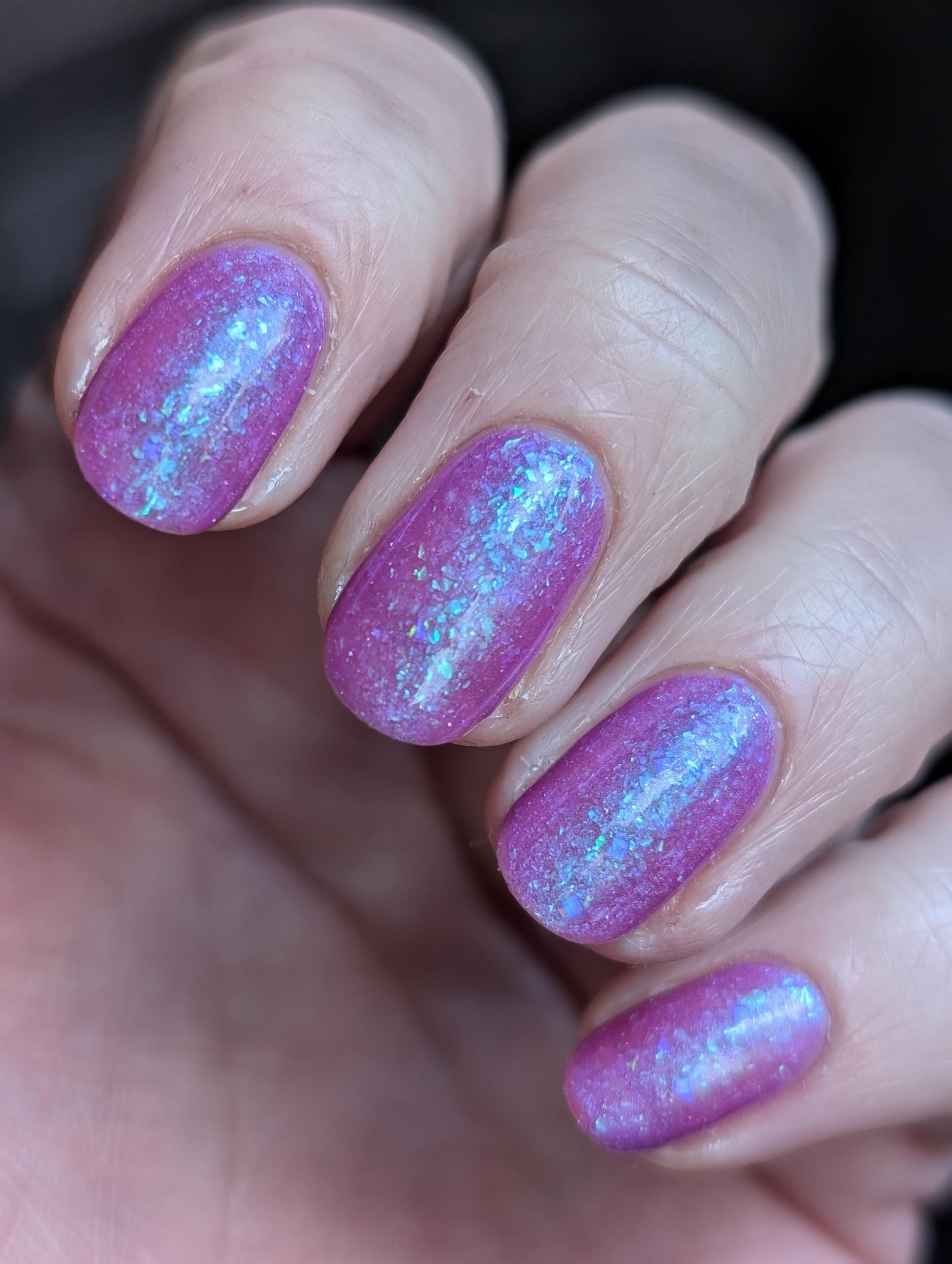 Closeup of nails painted in a lacquer that is bright pinkish purple sparkling with aqua shimmer and packed with iridescent flakes.