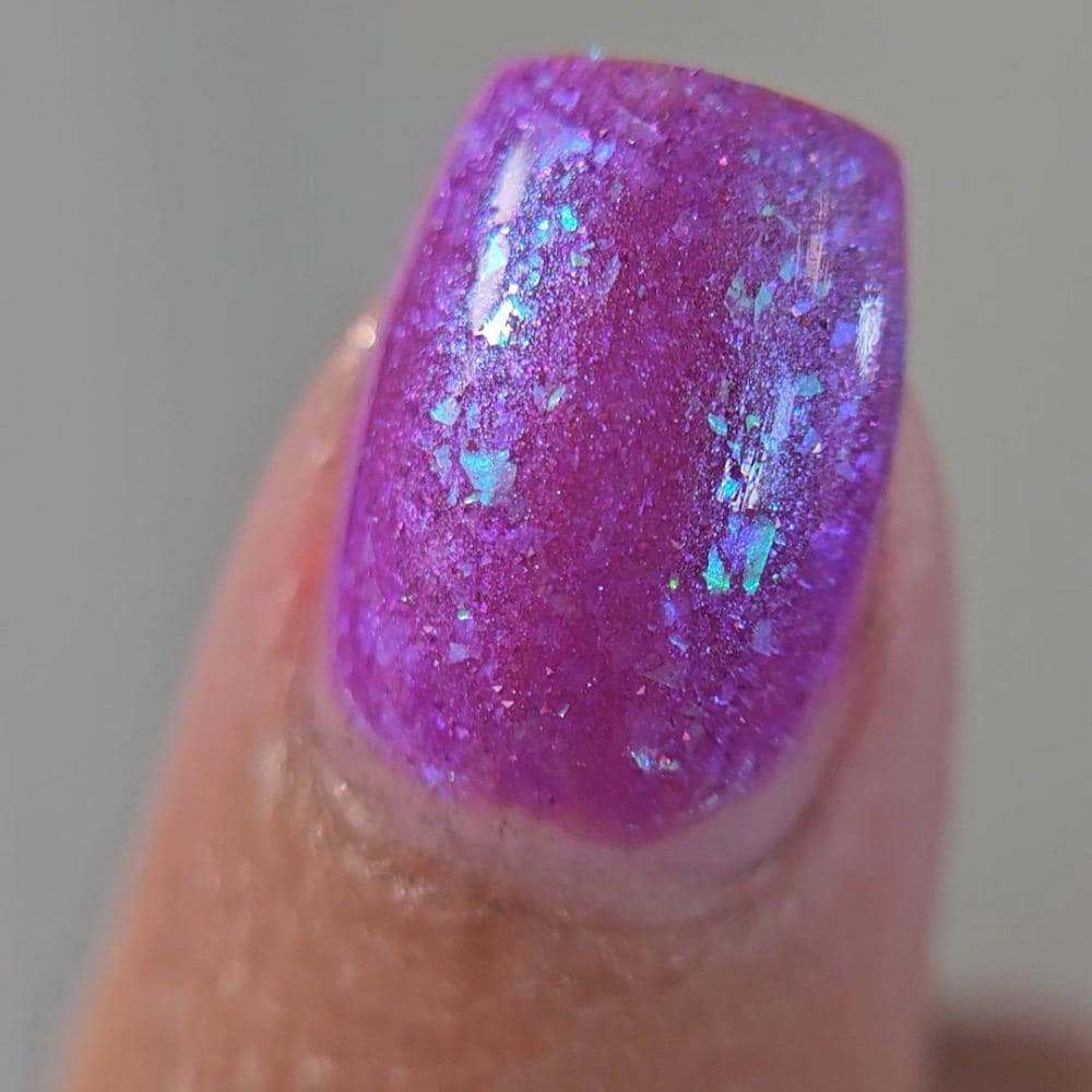 Closeup of  thumb painted in a lacquer that is bright pinkish purple sparkling with aqua shimmer and packed with iridescent flakes.