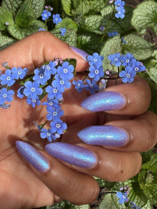 Hand with blue glittery nails holding blue flowers against a green leafy background
